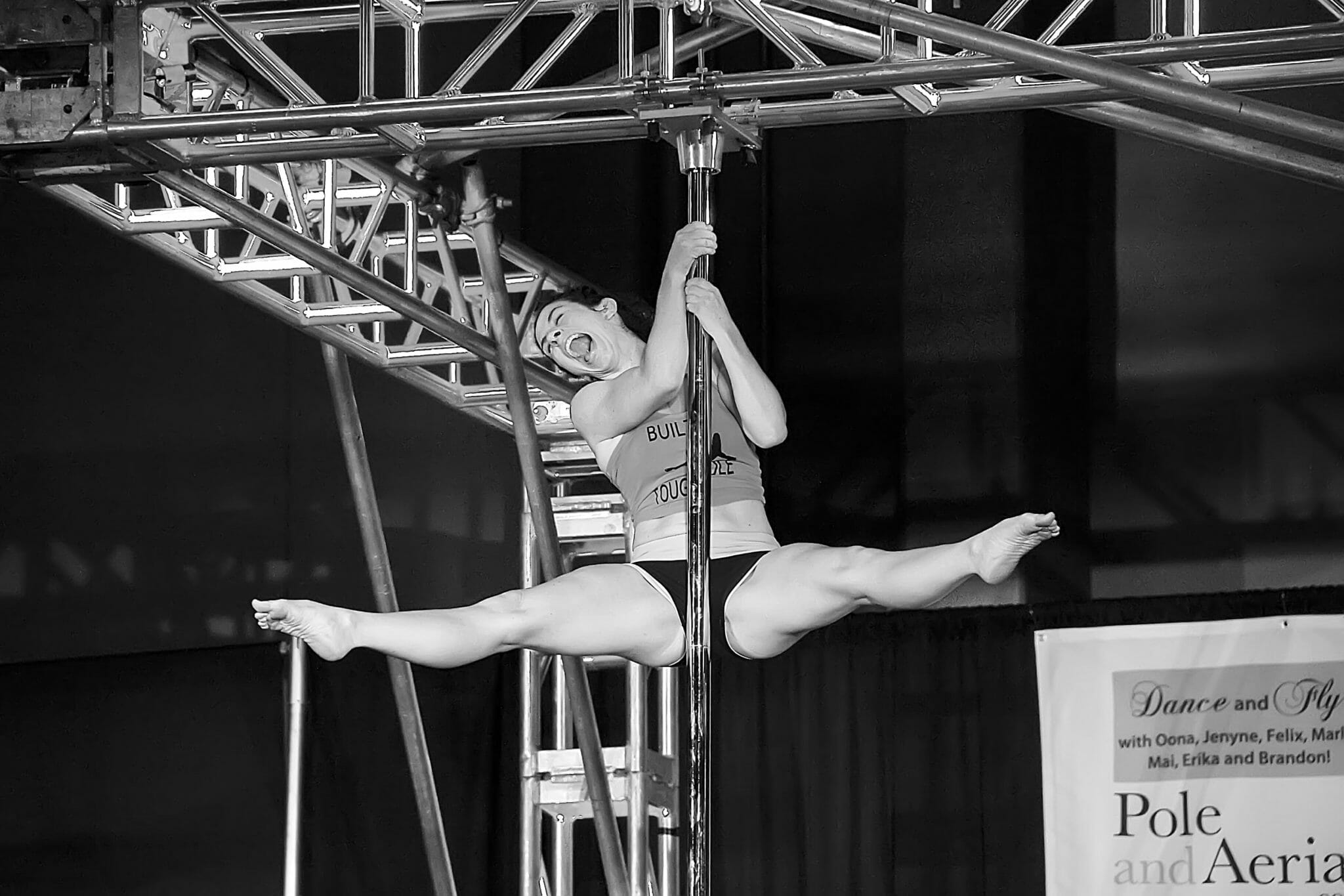 Pole dancer at the top of the pole in straddle, head-cocked as if yelling or shouting.