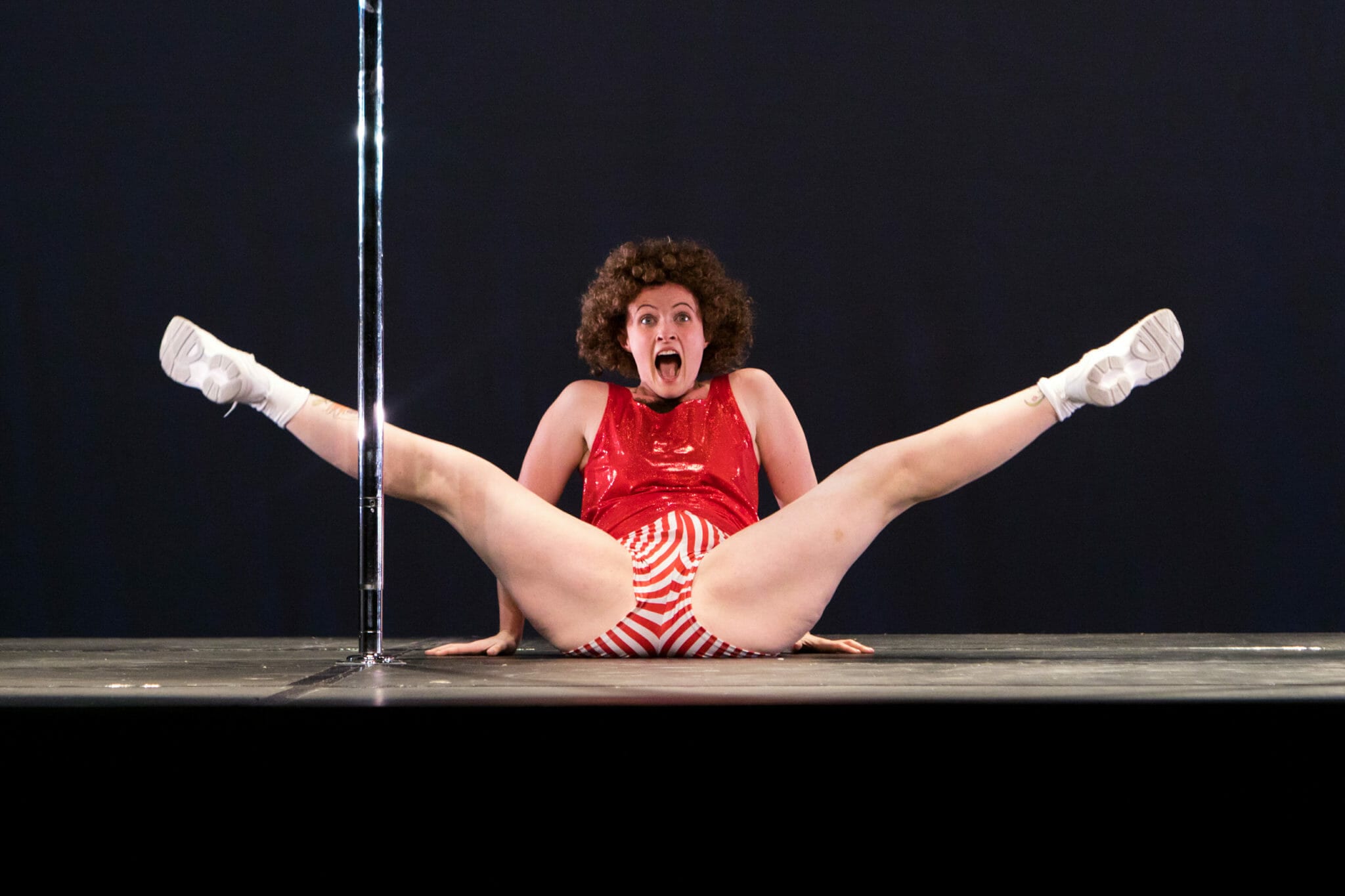 The Pole Comedian sits in straddle behind the pole during a performance.