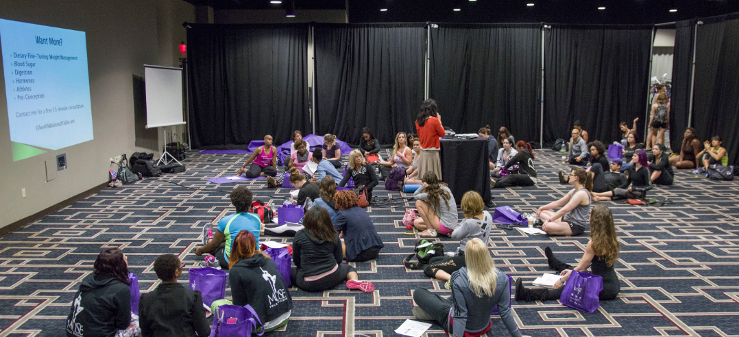 Attendees sit on the floor in a workshop room while speaker provides information via microphone