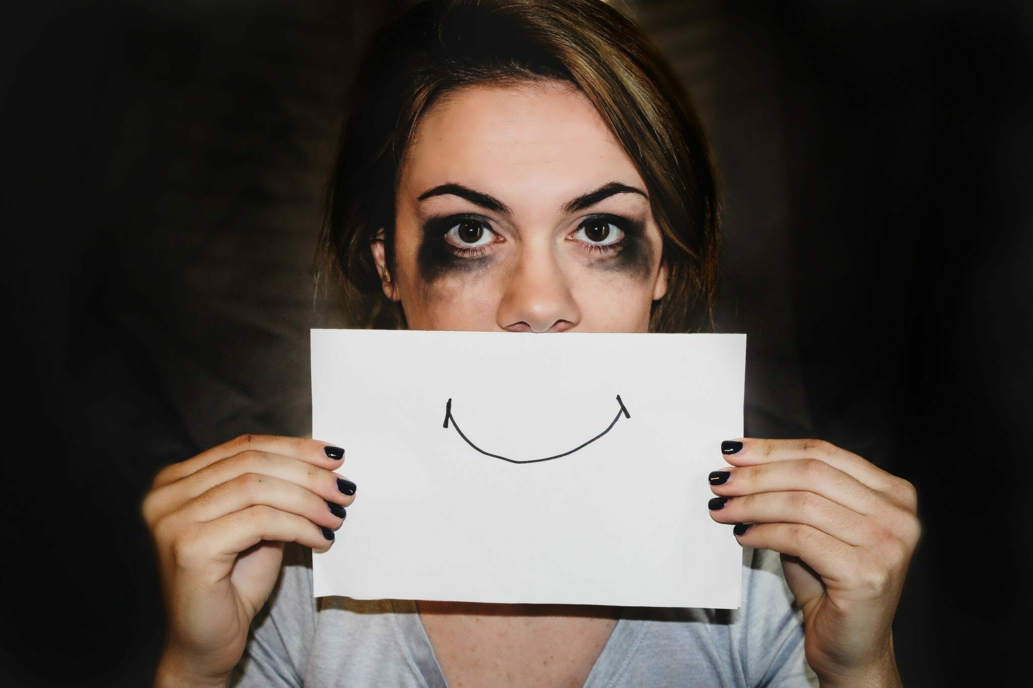 Individual stands with extremely smudged mascara while holding a half-sheet of paper with a smiley-face covering the lower portion of the face.