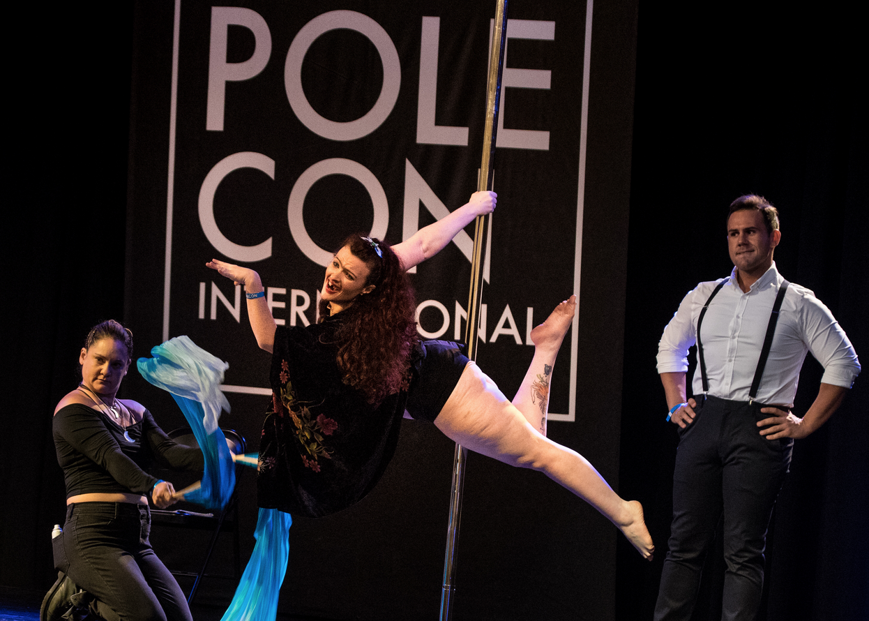 Three people on stage, one working silk fans, one in superman on the pole facing the audience, the third stands with hands on hips looking apprehensively at the pole dancer.