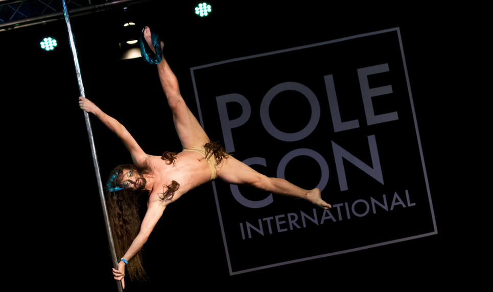 Male presenting Pole dancer executes an iron-x while wearing strategically glued long hair to cover groin and nipples.