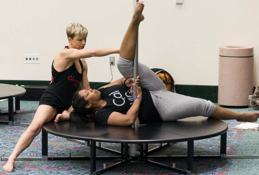 Instructor assists a student with leg positioning on an XPole stage during a workshop.