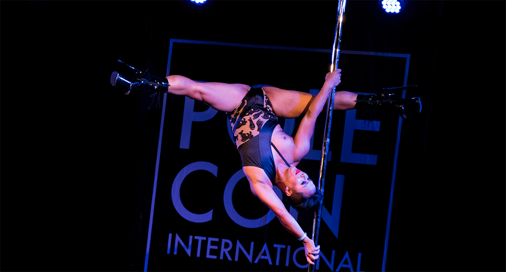 Pole dancer performs a split grip Ayesha on stage.
