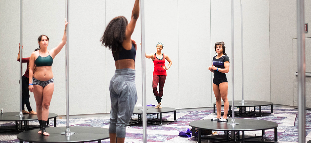 Students watch an instructor from X-pole stages scattered around a workshop room.