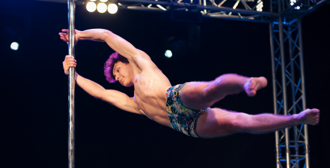 Male presenting pole dancer swings horizontally in cup grip.